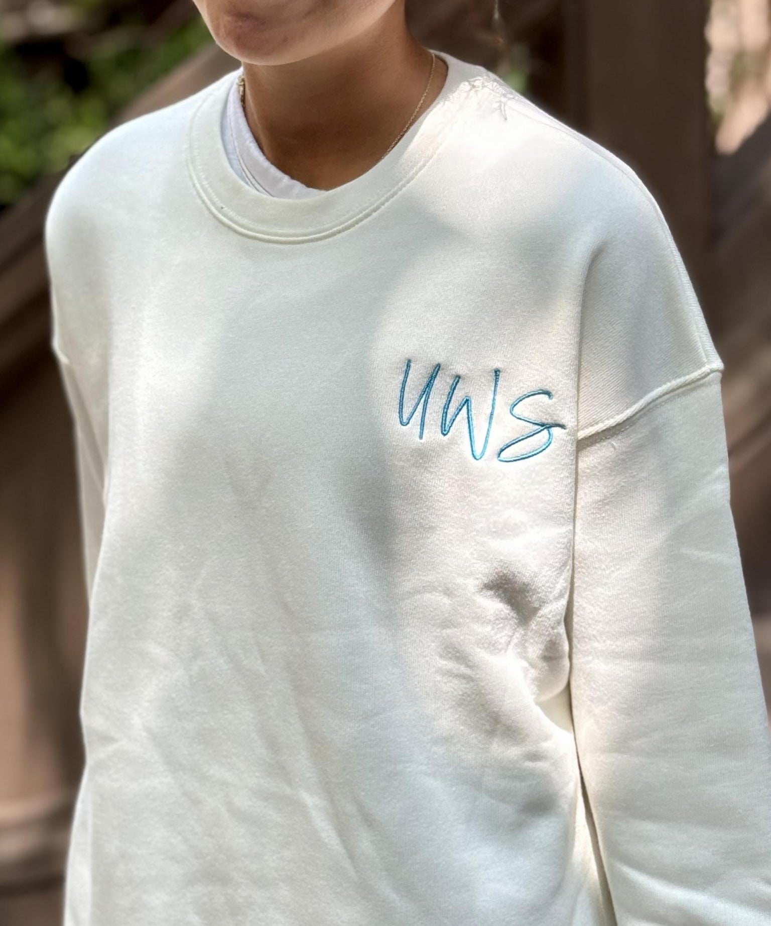 A close-up of a person wearing a cream-colored crewneck sweatshirt with the letters 'UWS' - upper west side -embroidered in light blue on the left chest, set against a blurred outdoor background.